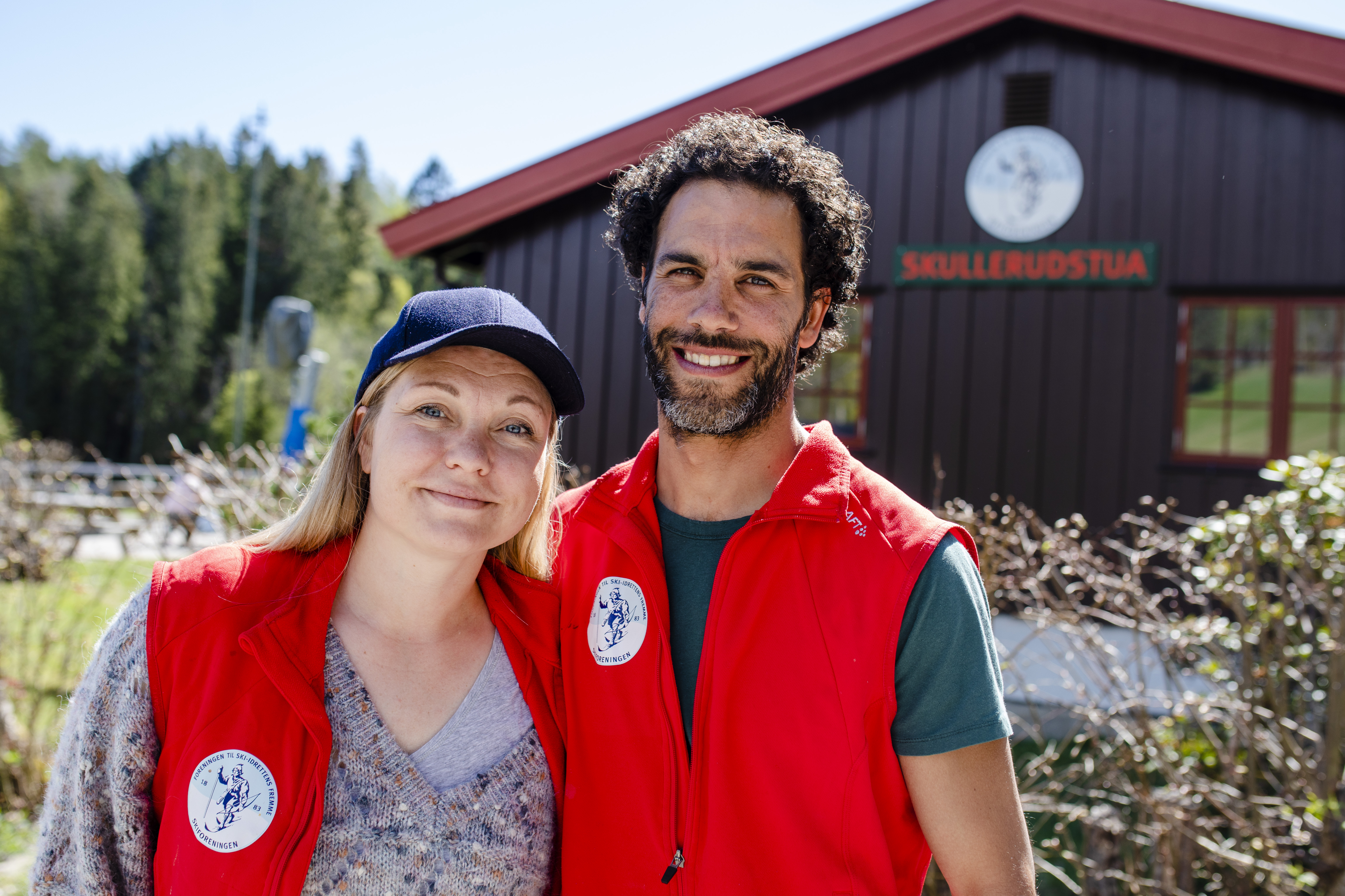To smilende verter i røde Skiforeningen-vester foran Skullerudstua en solrik dag